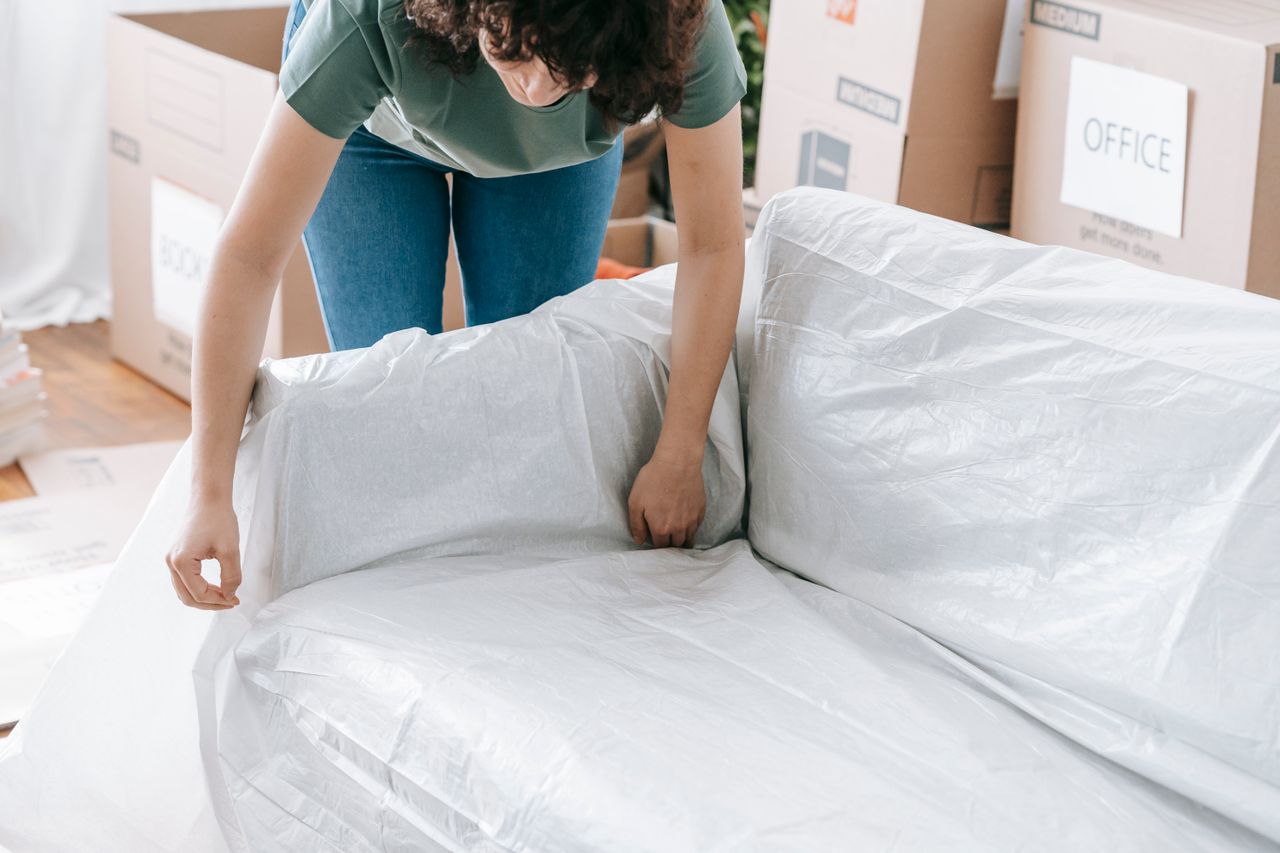 our-story A woman carefully covers a sofa with protective material while packing boxes in a bright room.